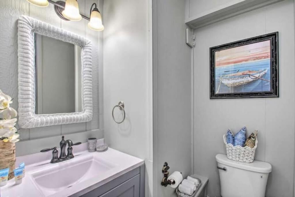 A bathroom with a white sink, a mirror, a framed painting of a boat, and a toilet with a basket of towels.
