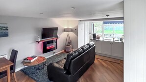 Ein Wohnzimmer mit einer schwarzen Ledersofa, einem Flachbildfernseher über einem Kamin, einem Esstisch mit Stühlen und Blick ins Freie durch ein Fenster.