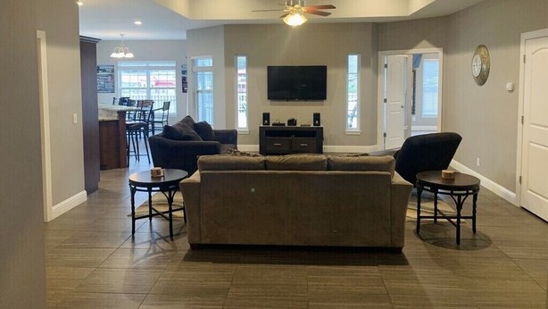 A spacious living room with a large sofa, two armchairs, a coffee table, and a ceiling fan. A flat-screen TV is mounted on the wall, and there is a dining area visible in the background.