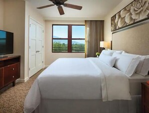 Relax in a spacious room with a large bed, ceiling fan, and TV on a stand, offering a serene view of the surrounding greenery and mountains.