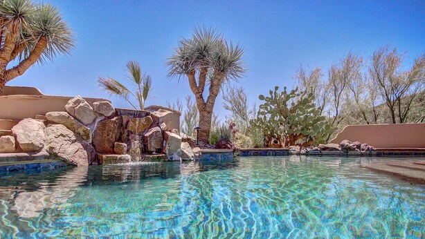 A clear pool with a rocky waterfall feature, surrounded by desert vegetation and a clear blue sky. - Fabulous Luxury Home on Quiet 2 acres (Tucson)