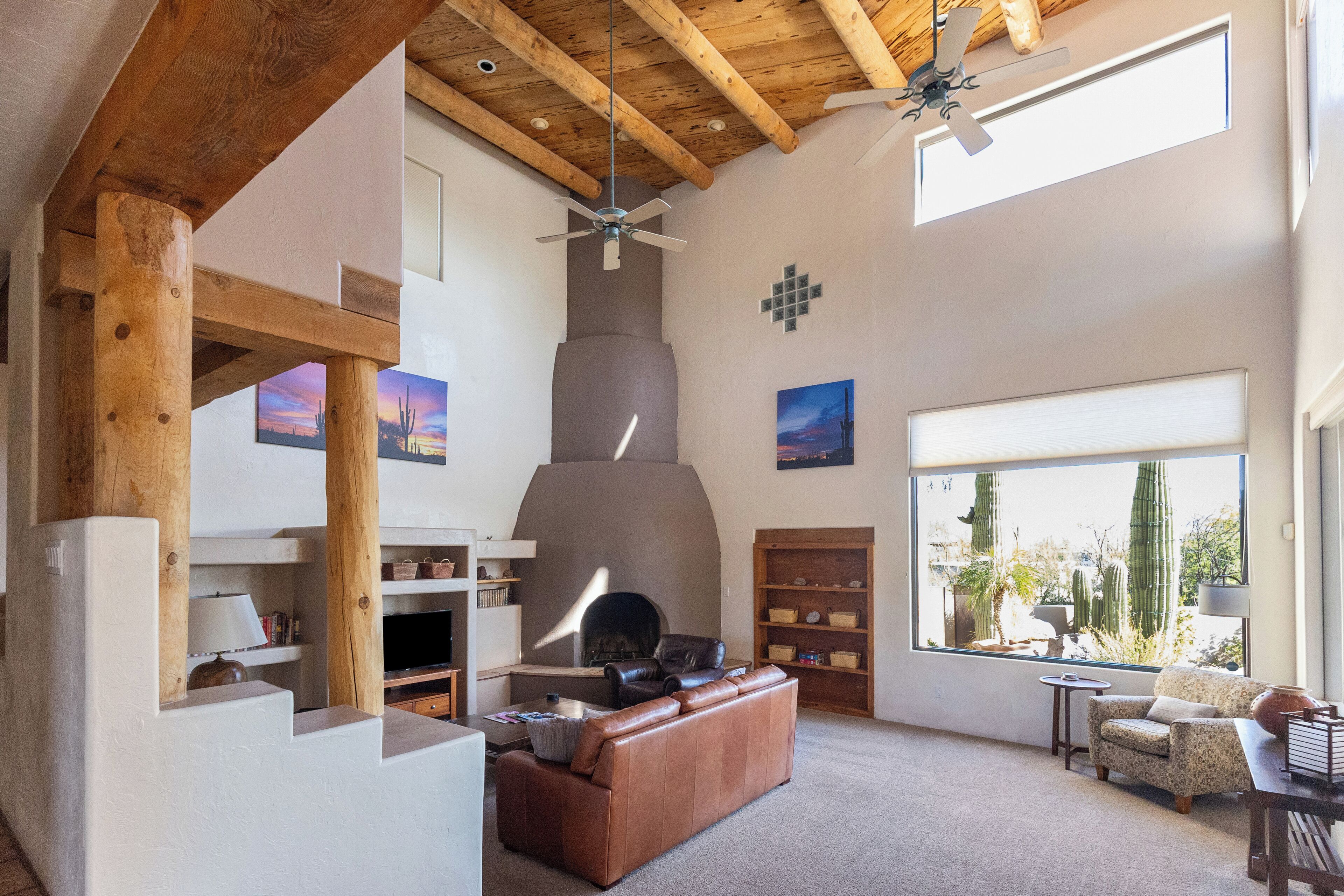Amplia sala de estar con chimenea, vigas de madera y ventanas grandes que ofrecen vista de cactus.