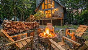 Een chalet met een vuurkuil, omringd door houten stoelen, een houtstapel en een terras.