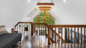 Een ruime loft slaapkamer met een schuine hemel, houten balken, een groot raam met uitzicht op groen, een donkergrijze bank en een trap met houten leuningen.
