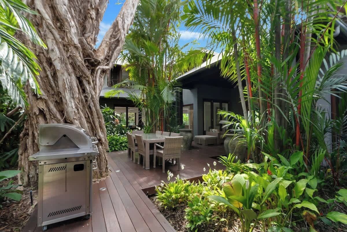 A wooden deck with outdoor furniture, a barbecue grill, and lush tropical plants.