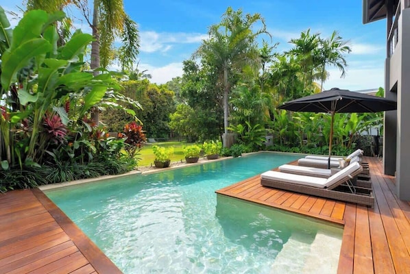 A pool with wooden deck and lounge chairs, surrounded by lush tropical plants.
