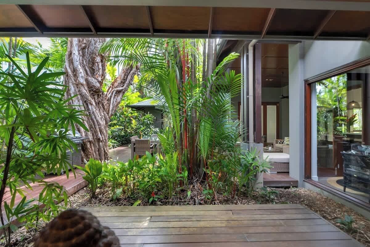 A modern house with a wooden deck, lush greenery, and a glass door leading to a patio area.