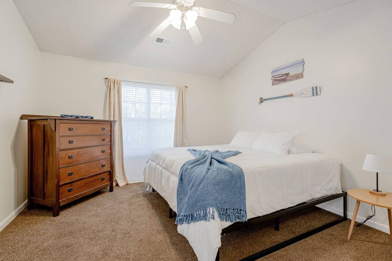 A bedroom with a bed, a dresser, a ceiling fan, a window with curtains, and a small table with a lamp.