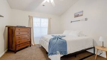 A bedroom with a bed, a dresser, a ceiling fan, a window with curtains, and a small table with a lamp.