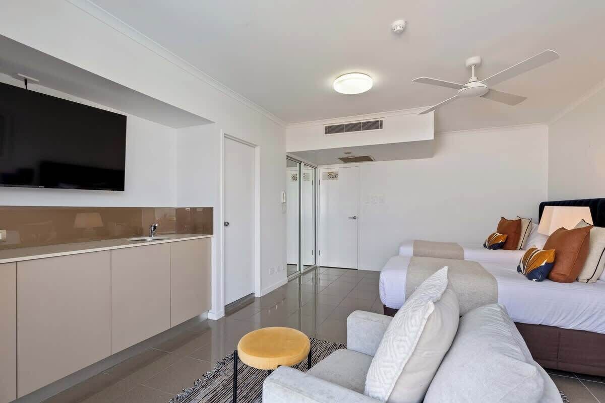 A modern hotel room with a flat-screen TV, a ceiling fan, a sofa, a bed with pillows, and a small round stool.