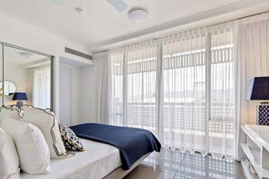 Une chambre d’hôtel avec un lit, une table de chevet avec une lampe, une grande fenêtre avec des rideaux légers et un ventilateur de plafond.