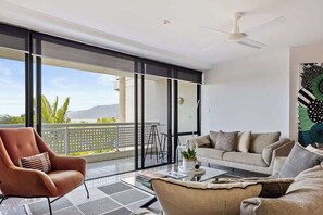 A modern living room with a sofa, armchair, and coffee table, large sliding glass doors to a balcony, and a ceiling fan.