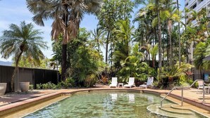 A clear pool with lounge chairs, surrounded by palm trees and a building in the background.