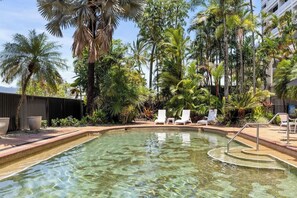 Una piscina cristallina con lettini prendisole, circondata da palme e un edificio sullo sfondo.