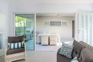 A modern living room with a sofa, a wicker chair, a dining table with chairs, and a sliding glass door leading to an outdoor area.