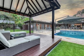 A covered outdoor area with a pool, a patio with furniture, and a house in the background.