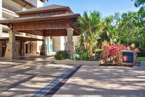 Ingresso di un hotel con una tettoia, colonne in pietra e un giardino curato con piante tropicali.