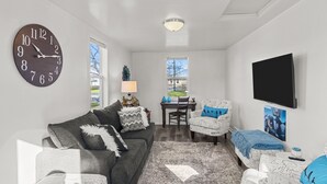 A living room with a large clock, a television, and a dining area with a table and chairs. - Charming Cottage (Machesney Park)