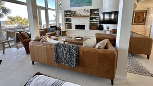 A spacious living room with a large window overlooking the ocean, a fireplace, and a flat-screen TV mounted on the wall.