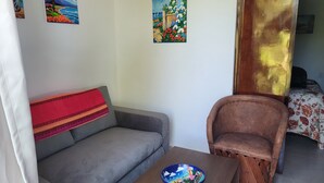 A living room with a grey sofa, a wooden chair, a small table with a blue bowl, and three paintings on the wall. - Tiny Condo PB 5 at Vista Encantada! 1 Bedroom, kitchen, pool, incredible views!  (El Divisadero)