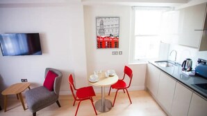 Cocina compacta con una mesa de comedor, sillas rojas, un taburete pequeño y un televisor de pantalla plana montado en la pared.