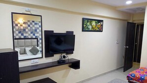 A hotel room with a bed, a TV mounted on the wall, a small table, a bench, a colorful bedspread, a tiled wall, and a door.