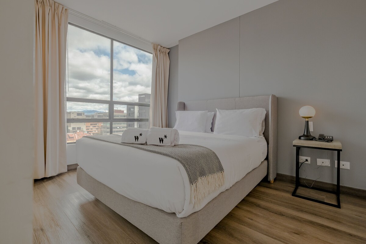 Una habitación de hotel moderna con una cama grande, una mesita de noche con lámpara y vistas de la ciudad a través de un ventanal amplio.