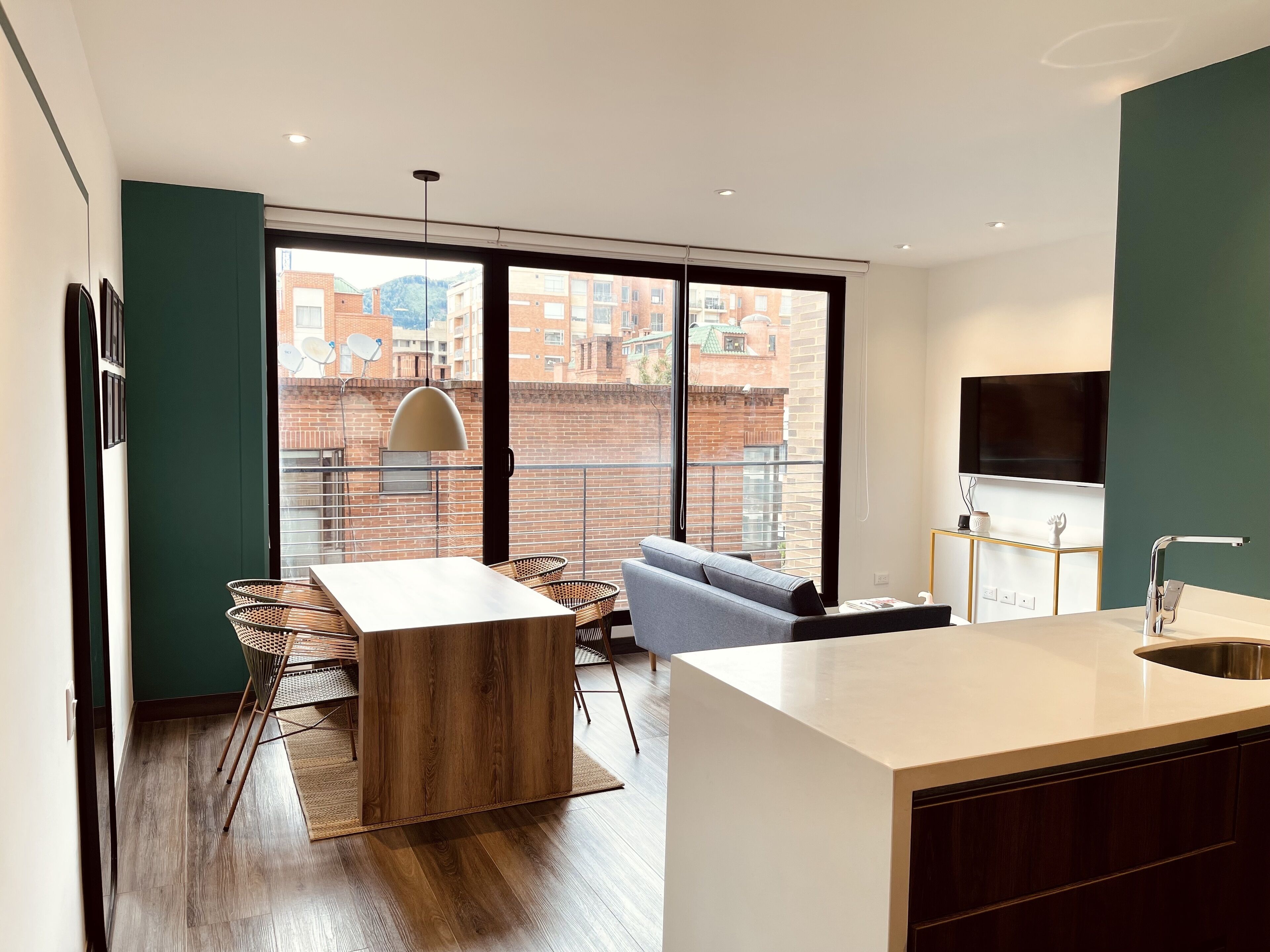 A modern kitchen with a large island, a dining area with a table and chairs, and a view of a brick building through large windows.