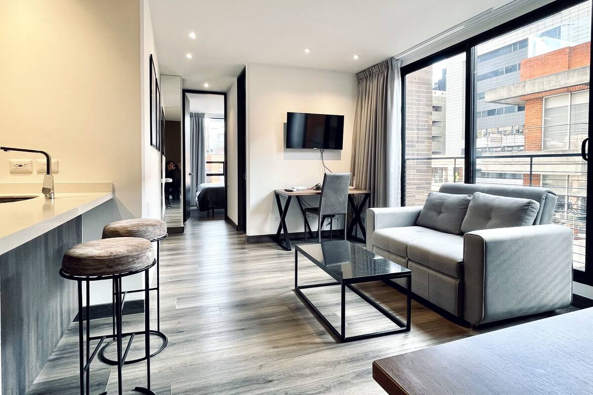 Una habitación de hotel moderna con sofá, una mesa de centro, televisor y balcón con vista a la ciudad.