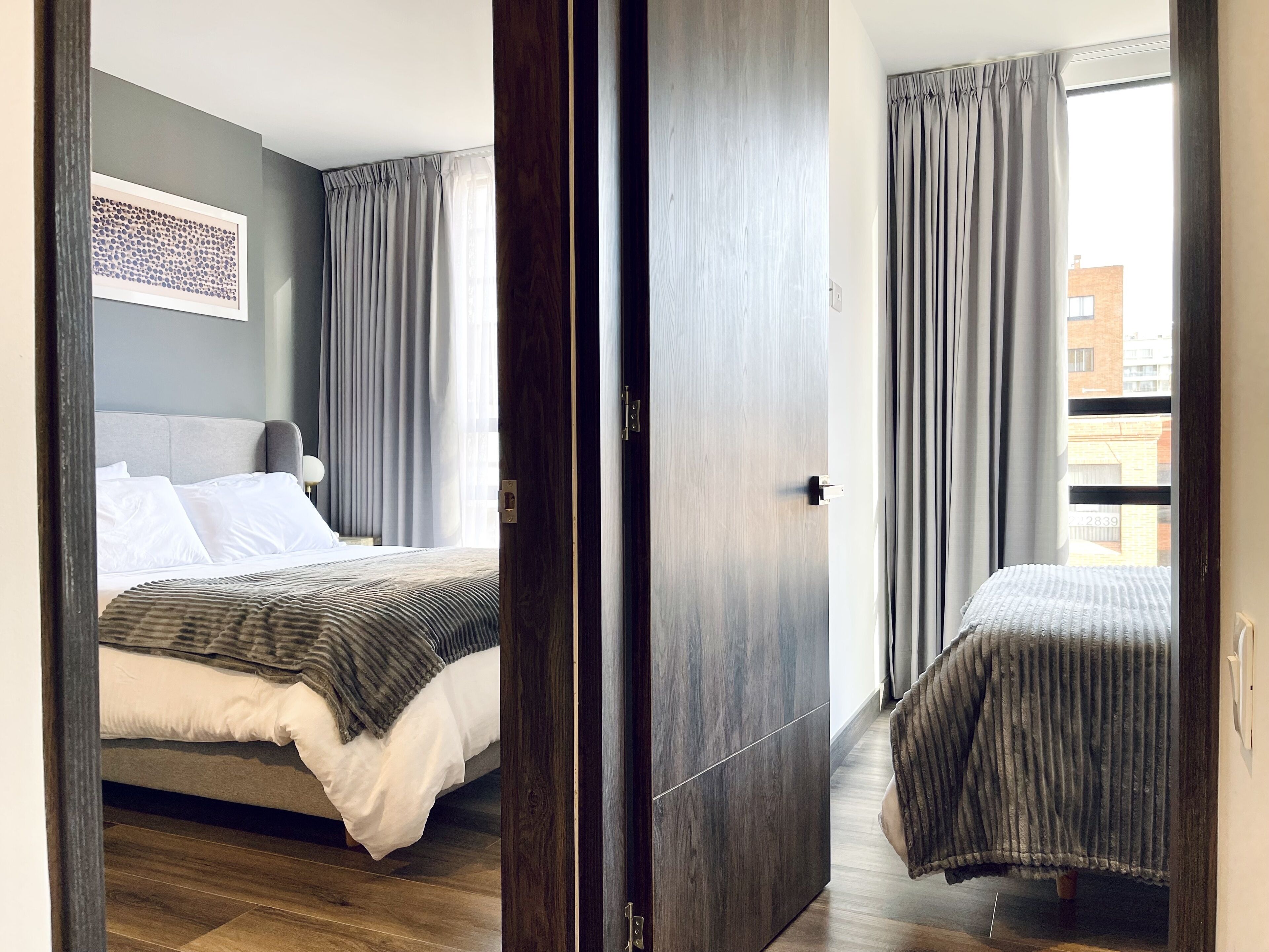 A modern hotel room with a bed, a bedside table, a large window with curtains, and a view of the city.