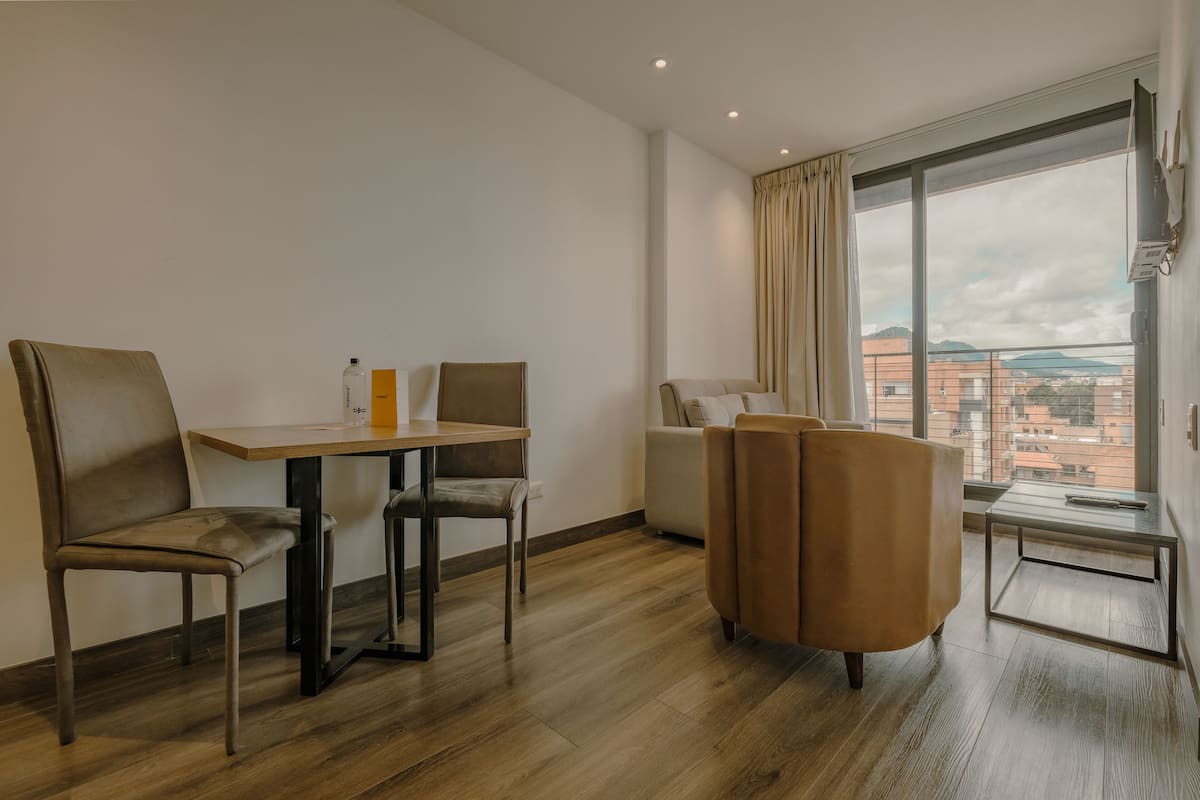 Habitación de hotel moderna con suelo de madera, una mesa de comedor con sillas, un sofá y un ventanal con vistas a la ciudad.