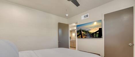 A hotel room with a bed, a ceiling fan, a painting of a sunset, and a view into the kitchen area.