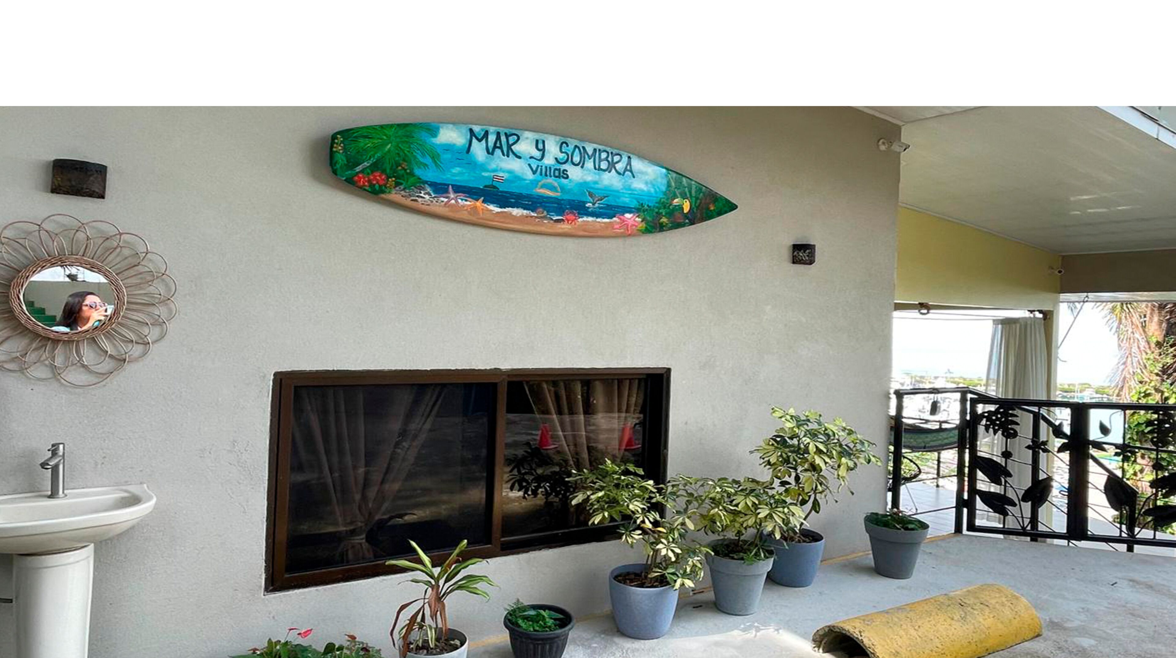 A room with a large window, a wall-mounted mirror, a potted plant arrangement, and a sign reading 'MAR y SOMBRA Villas'.