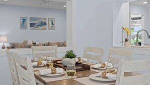 Ein Essbereich mit einem Holztisch, gedeckt für vier Personen, weißen Stühlen und Blick auf ein Wohnzimmer mit einer Couch und Kunstwerken an der Wand.