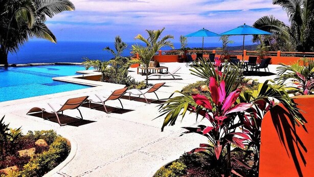 A poolside area with lounge chairs and umbrellas, surrounded by tropical plants and palm trees, overlooking a clear blue sky and ocean. - 1 Bedroom Condo with Incredible Ocean View in Nayarit Jungle #404 (El Divisadero)