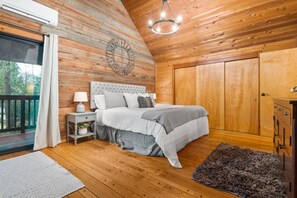 A bedroom with a wooden ceiling, a bed with grey bedding, a nightstand with a lamp, and a large wooden wardrobe.