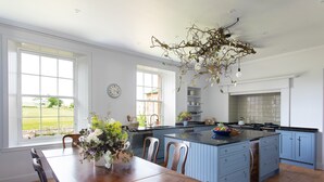A spacious kitchen with a large island, wooden dining table, and chairs. There are windows providing natural light and a view of the outdoors. - Idyllic setting for large family get-togethers, groups of friends, and corporate retreats. (Haddington)