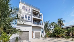 A modern two-story building with a balcony on the upper floor, surrounded by palm trees and a sandy ground.