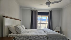 Quarto com cama, mesa de cabeceira, ventilador de teto e janela com cortinas.