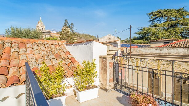 Una terraza en la azotea con baldosas de terracota, plantas en macetas, una reja metálica y una vista distante de una torre con campanas.