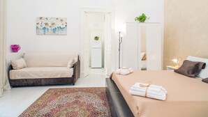 Habitación de hotel con cama, sofá, una mesita con lámpara, armario y puerta que da a otra habitación.