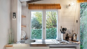 A kitchen with a double sink, wooden shelves, a hanging light, and a view of greenery outside. - Studio Apartment 'Little California' with Private Terrace, Wi-Fi and Air Conditioning (Cagnes-sur-Mer)
