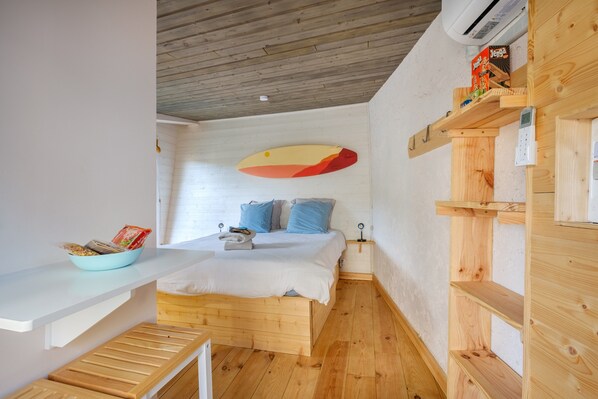 A room with a bed, a wooden bench, a shelf with a book, and a wall-mounted shelf with a box.