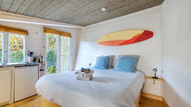 A bedroom with a bed, a bedside table, a wall-mounted surfboard, a window with curtains, and a small kitchen area. - Studio Apartment 'Little California' with Private Terrace, Wi-Fi and Air Conditioning (Cagnes-sur-Mer)