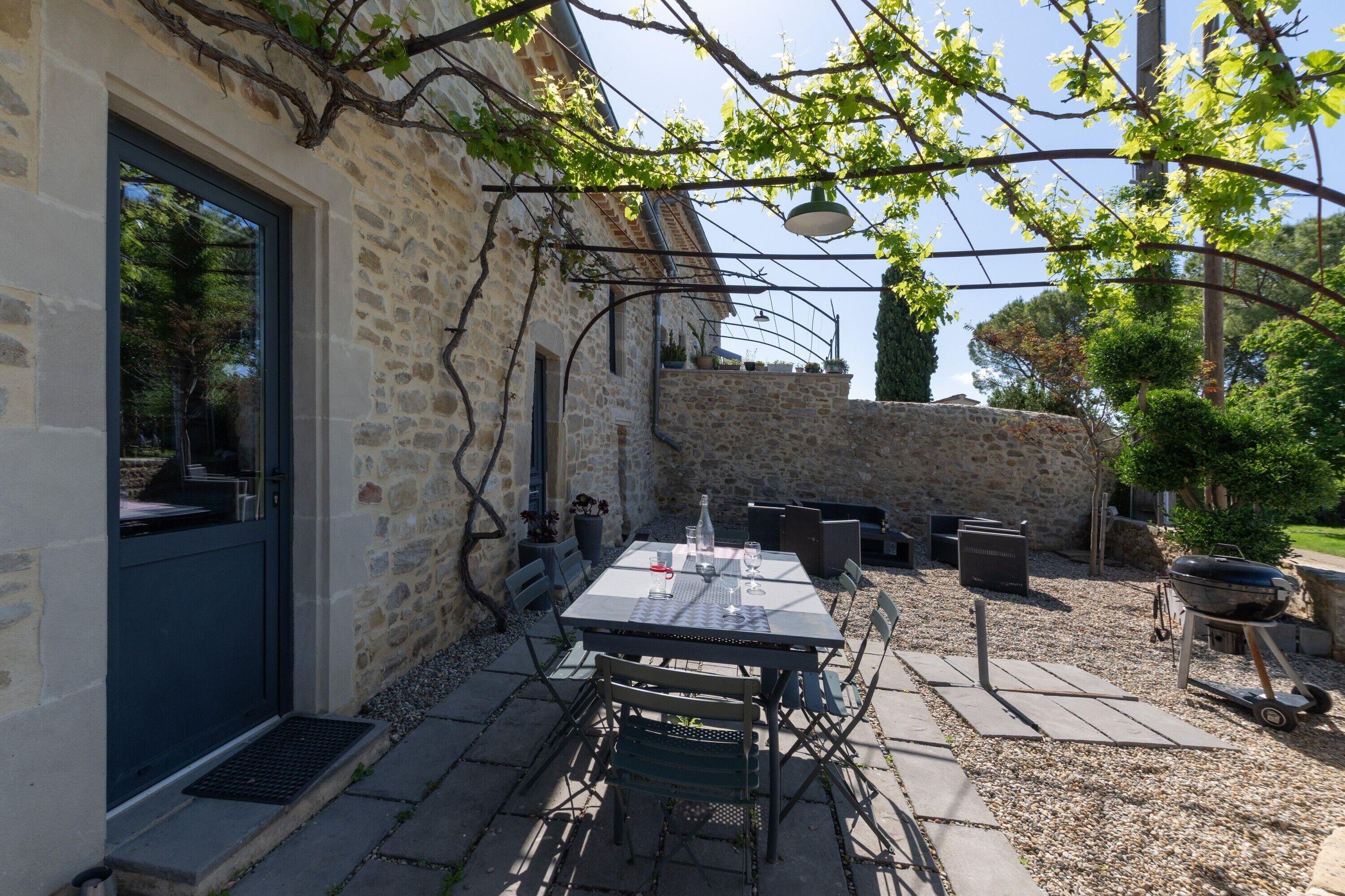 A stone building with a dark door, outdoor seating with tables and chairs, a barbecue grill, and potted plants.