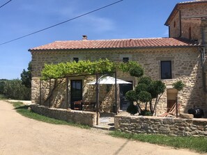 A stone building with a red-tiled roof, a covered porch with a table and chairs, and a small garden with potted plants. - Holiday Home 'Gîte Les Vignes Au Mas Dujau' with Shared Pool, Wi-Fi and Air Conditioning (Saint Sauveur de Cruzières)