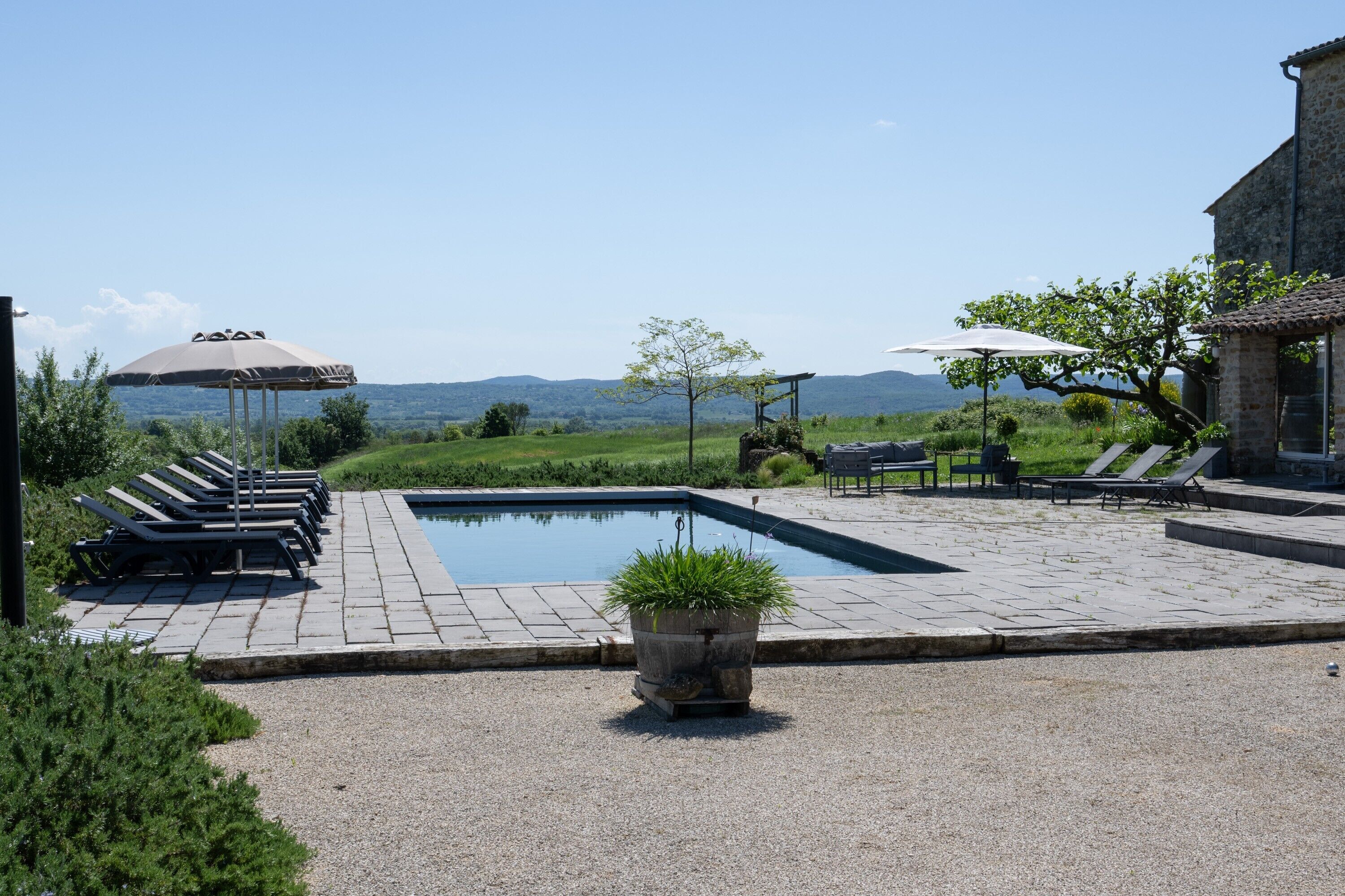 Zona de piscina con sillones de descanso y sombrillas, un edificio de piedra y vistas al paisaje.