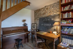 Una stanza con un pianoforte, una scrivania con una lampada, una libreria e un muro in pietra.
