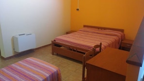 Une petite chambre d’hôtel composée d’une seule pièce, avec un lit, une table de chevet en bois, une petite table, une chaise et un radiateur fixé au mur.