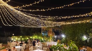 Un espace de restauration en plein air, agrémenté de guirlandes lumineuses suspendues, de tables dressées pour les clients et d’une végétation en arrière-plan.